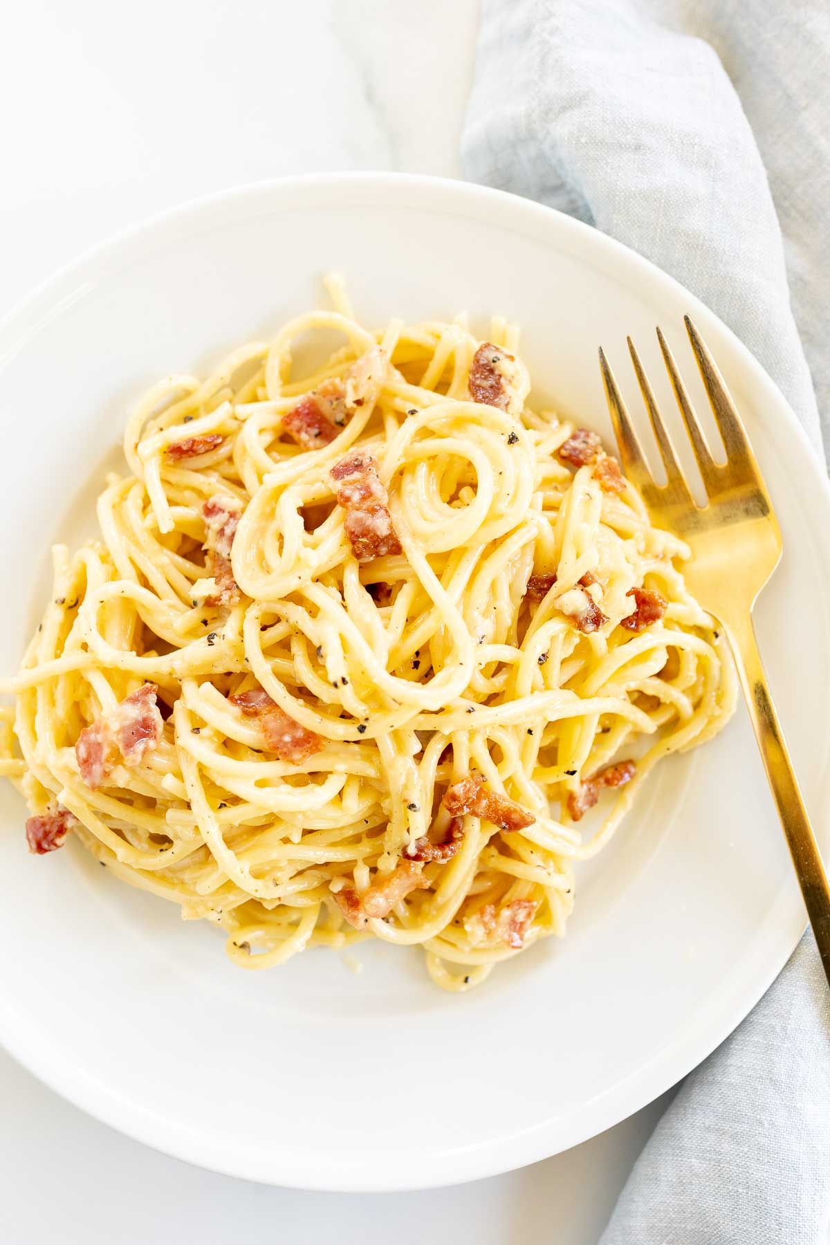 A white plate with a serving of spaghetti carbonara, gold fork to the side