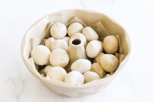 A bundt pan filled with raw dough balls, ready to become delicious monkey bread, rests on a white surface.