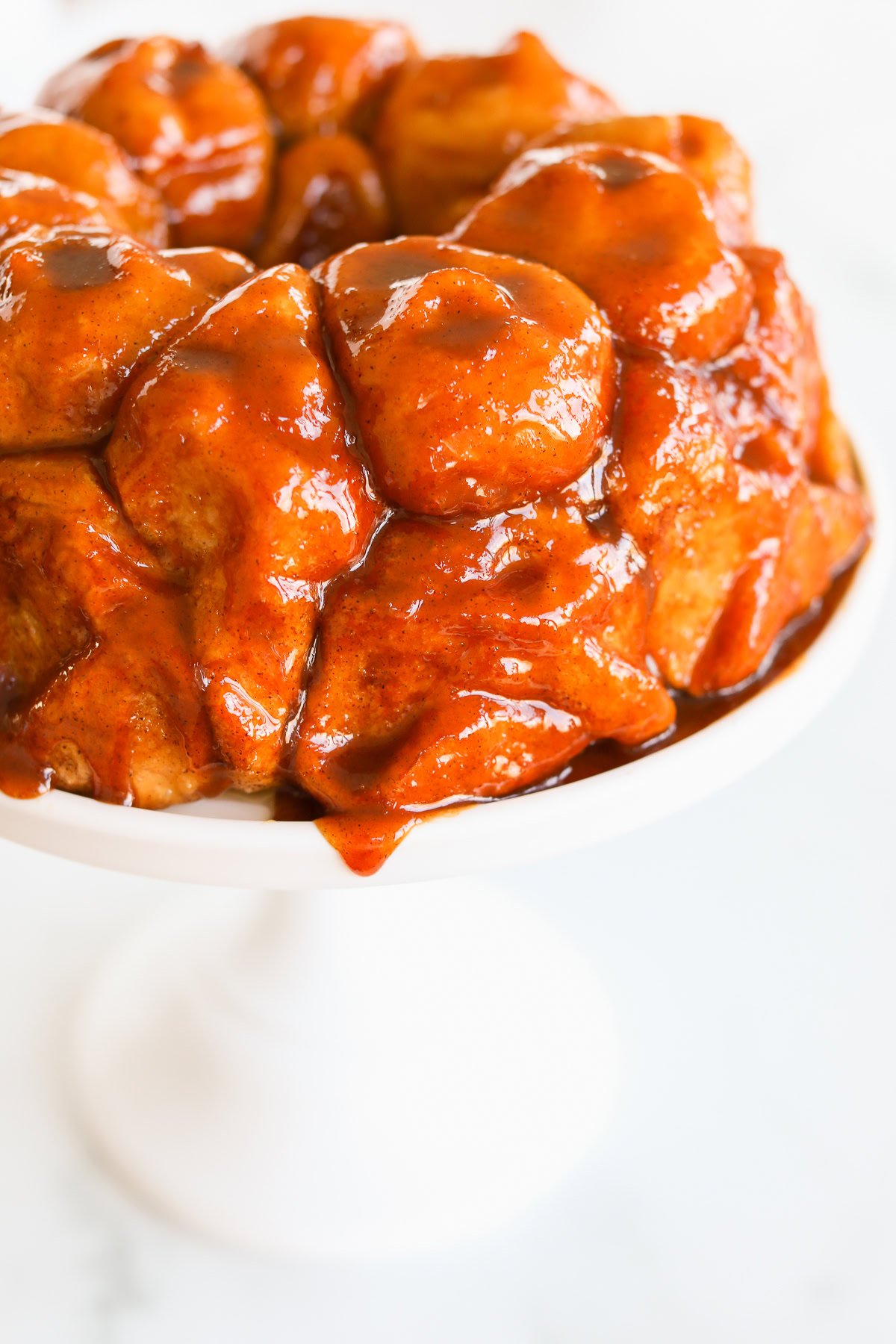 A close-up of a deliciously glazed monkey bread is artfully displayed on a pristine white cake stand, set against a light background.