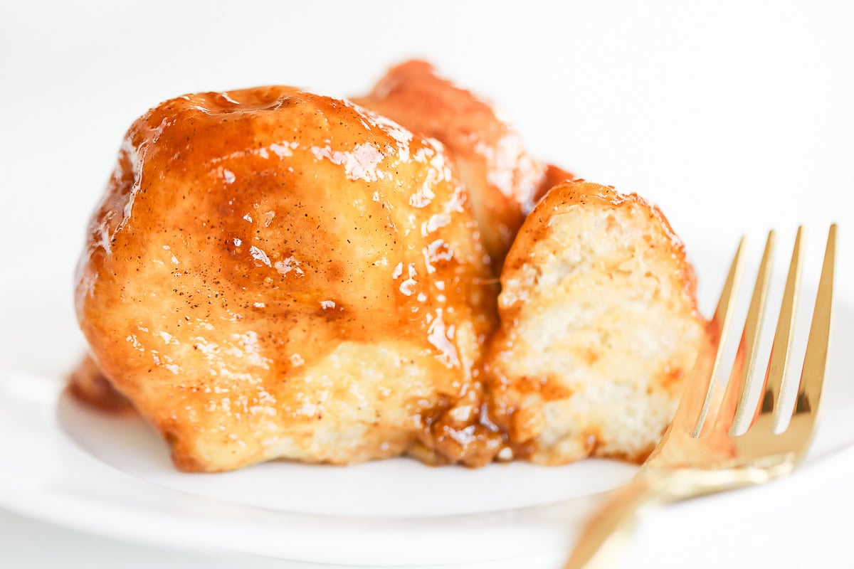 Close-up of a sticky, glazed dessert roll reminiscent of monkey bread on a white plate with a gold fork gleaming to the right.