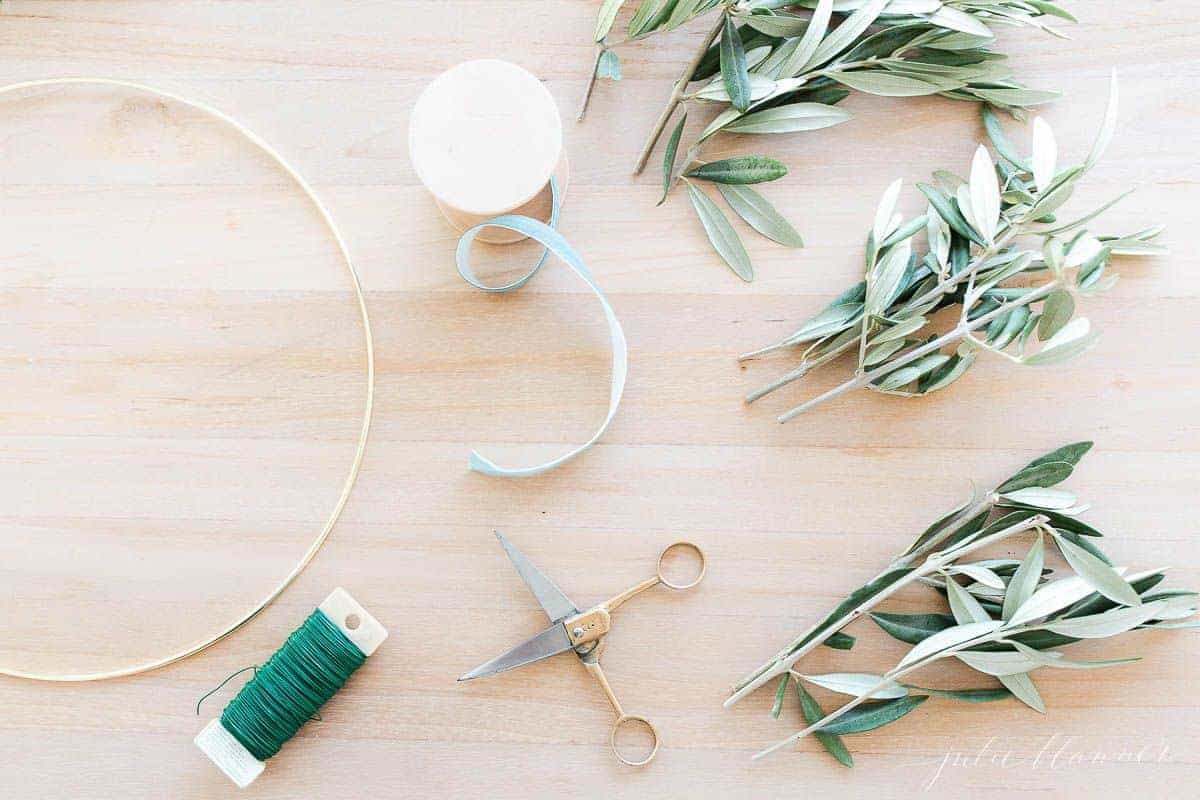 A wood table with a gold hoop. scissors, wire, ribbon and greenery laid out for making a wreath.