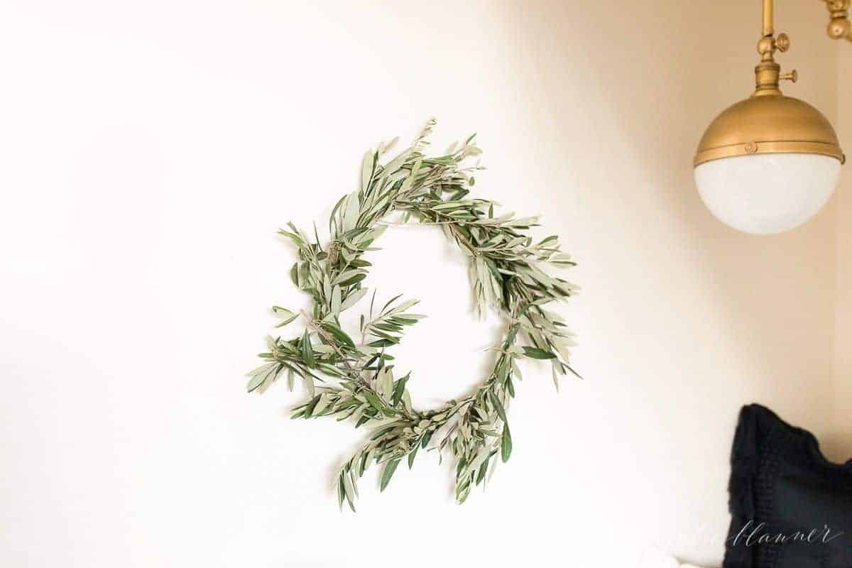 A white wall with a small DIY olive wreath hanging in front of a gold wall sconce.