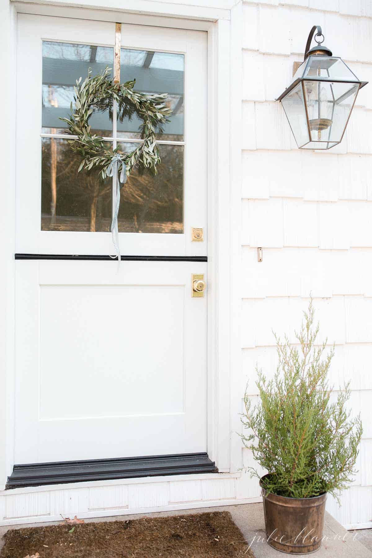 A small handmade olive wreath hangs on the top half of a white dutch door, with a small Christmas tree to the side. 