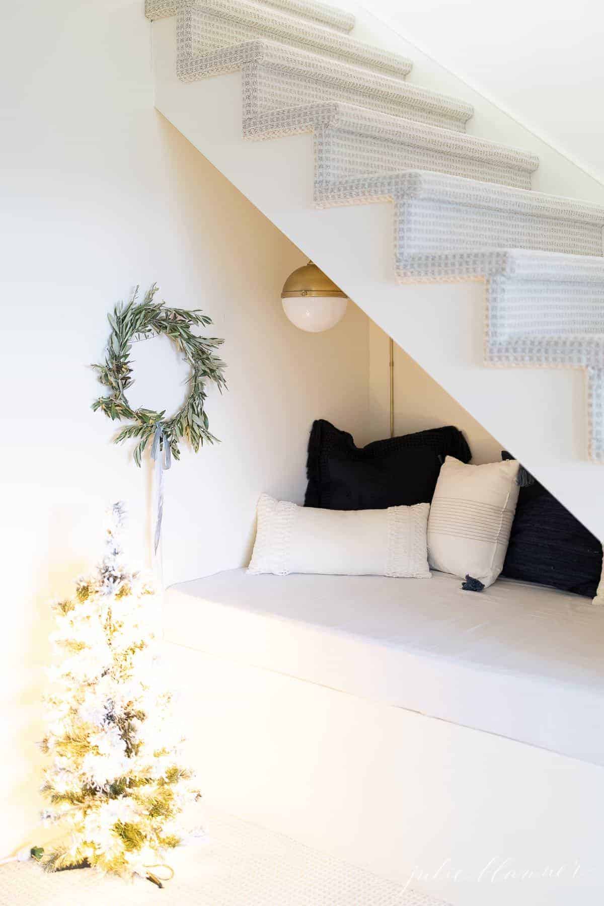 A DIY Christmas wreath hanging in a nook under the stairs piled with blue and white pillows.