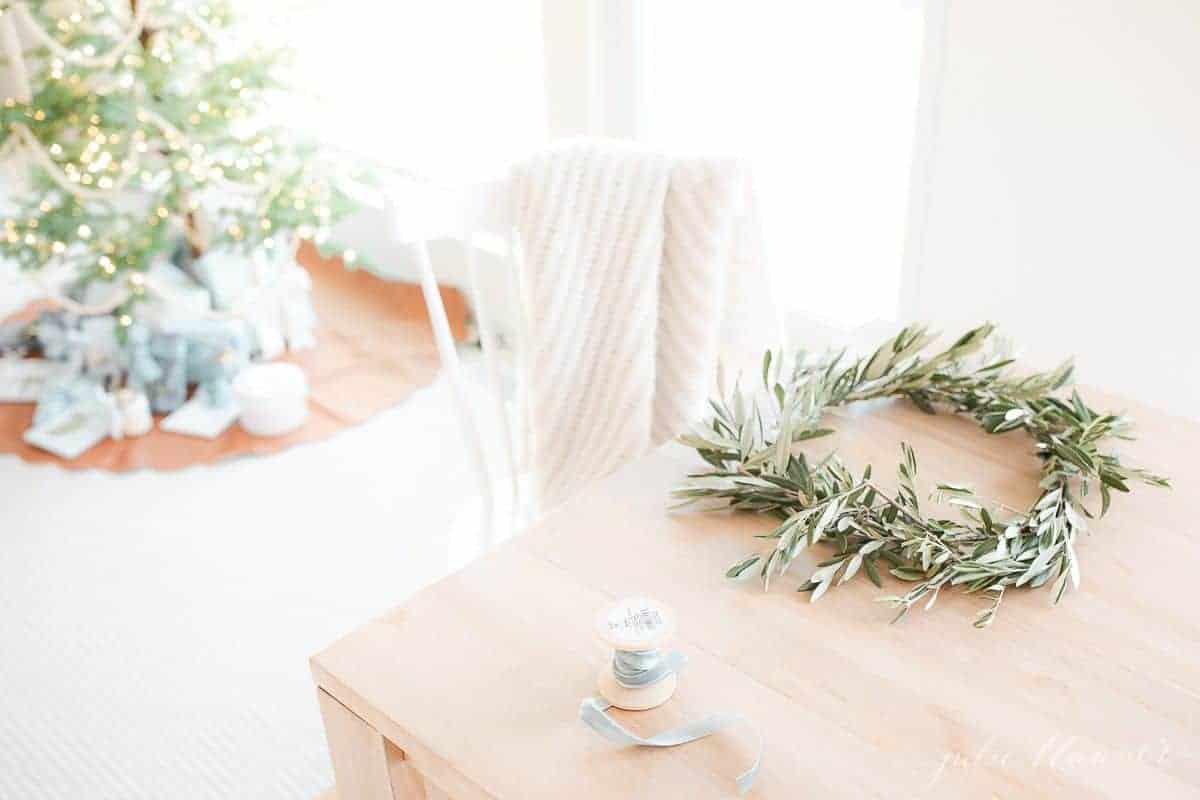 A small handmade olive wreath on a wooden table, christmas tree in background.