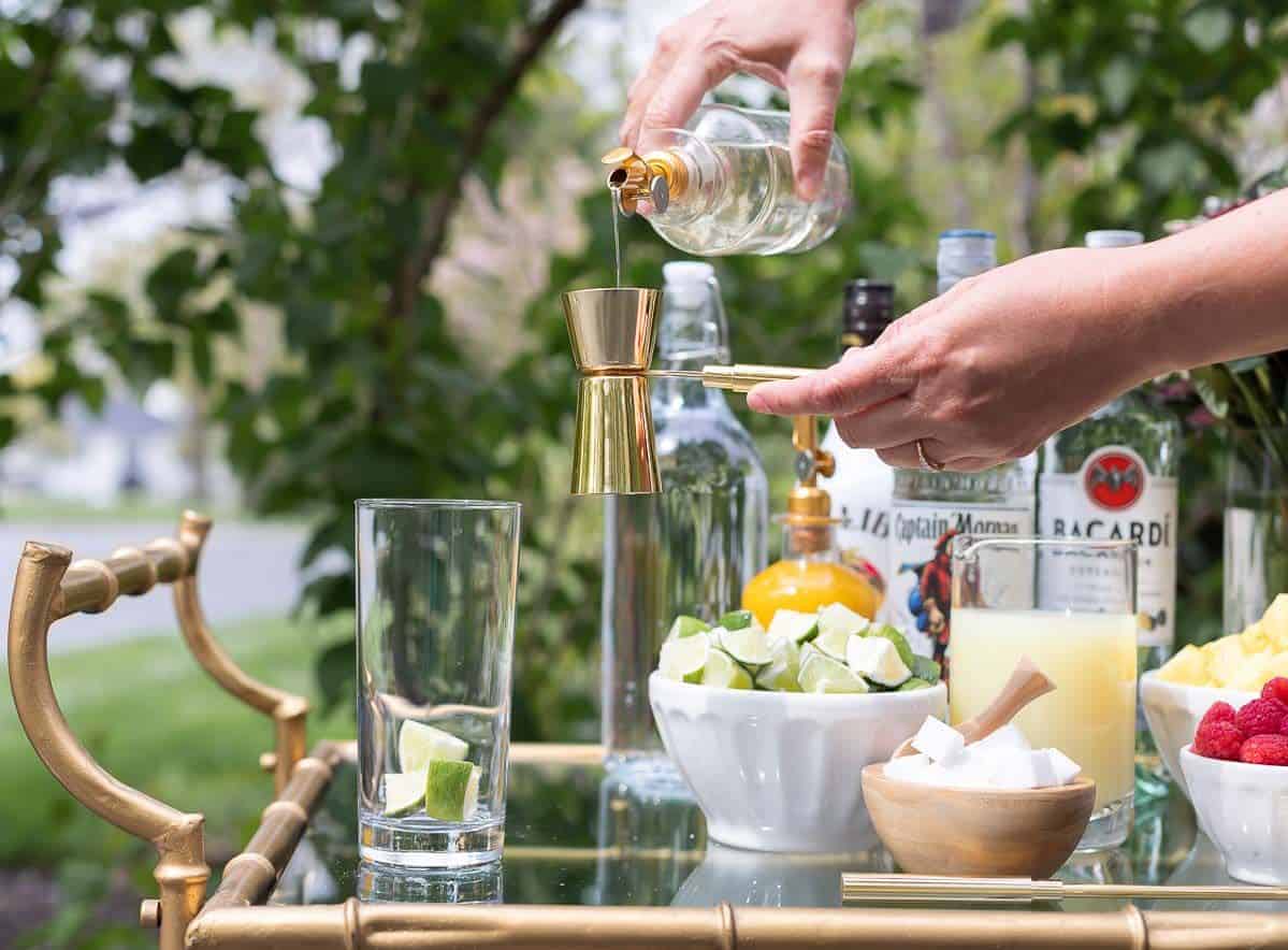 A mojito bar set up on a gold and glass bar cart, a hand reaching over to pour simple syrup in a glass.