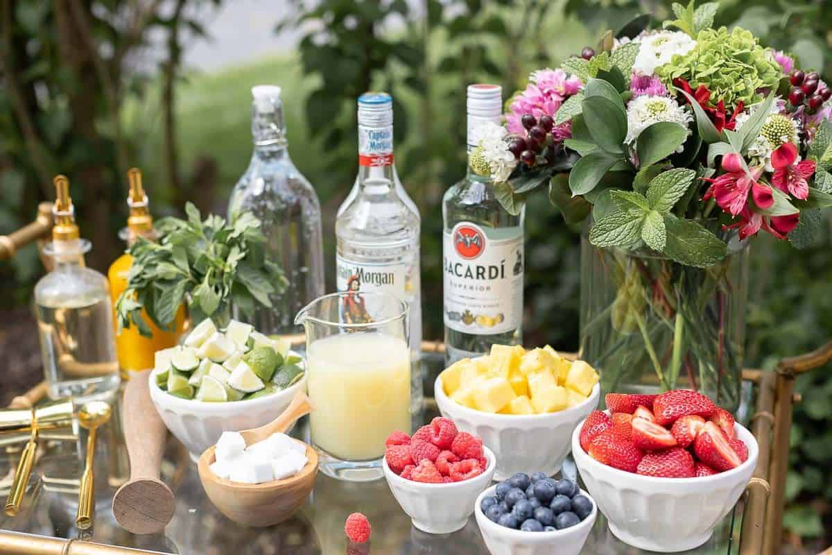 A glass and brass bar cart set up with a mojito bar outdoors, various fruits and limes in bowls beside alcohol bottles.