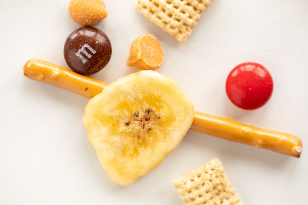 Dried banana chips, pretzels, candy and cereal on a white surface