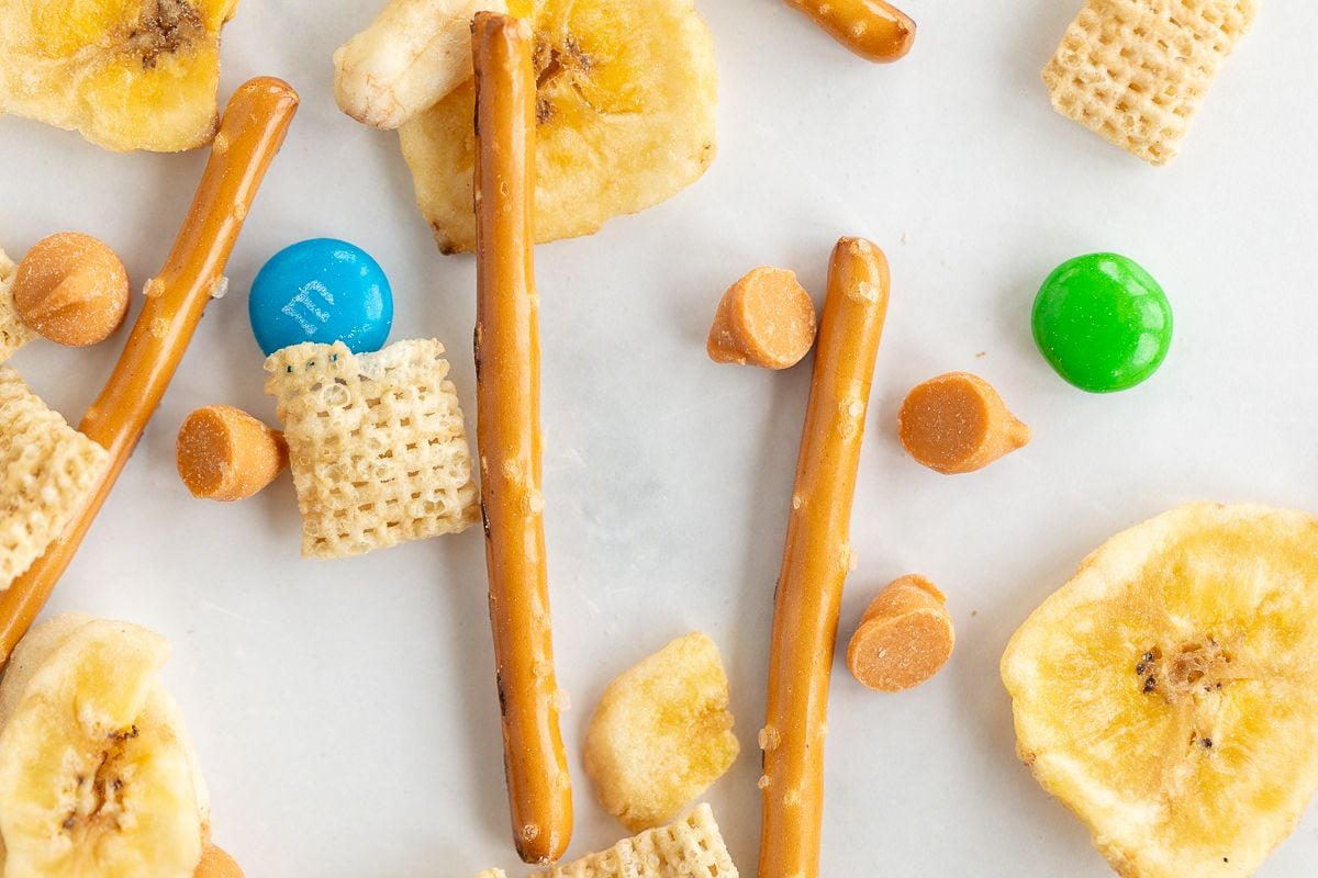 Dried banana chips, pretzels, candy and cereal spread out on a white surface