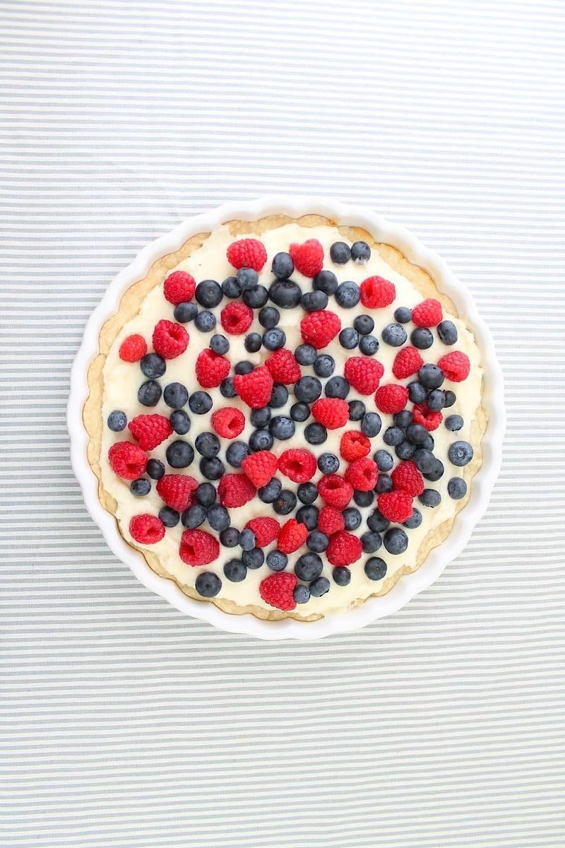 A round lemon berry tart, topped with fresh raspberries and blueberries, sits elegantly on a striped white and grey background.