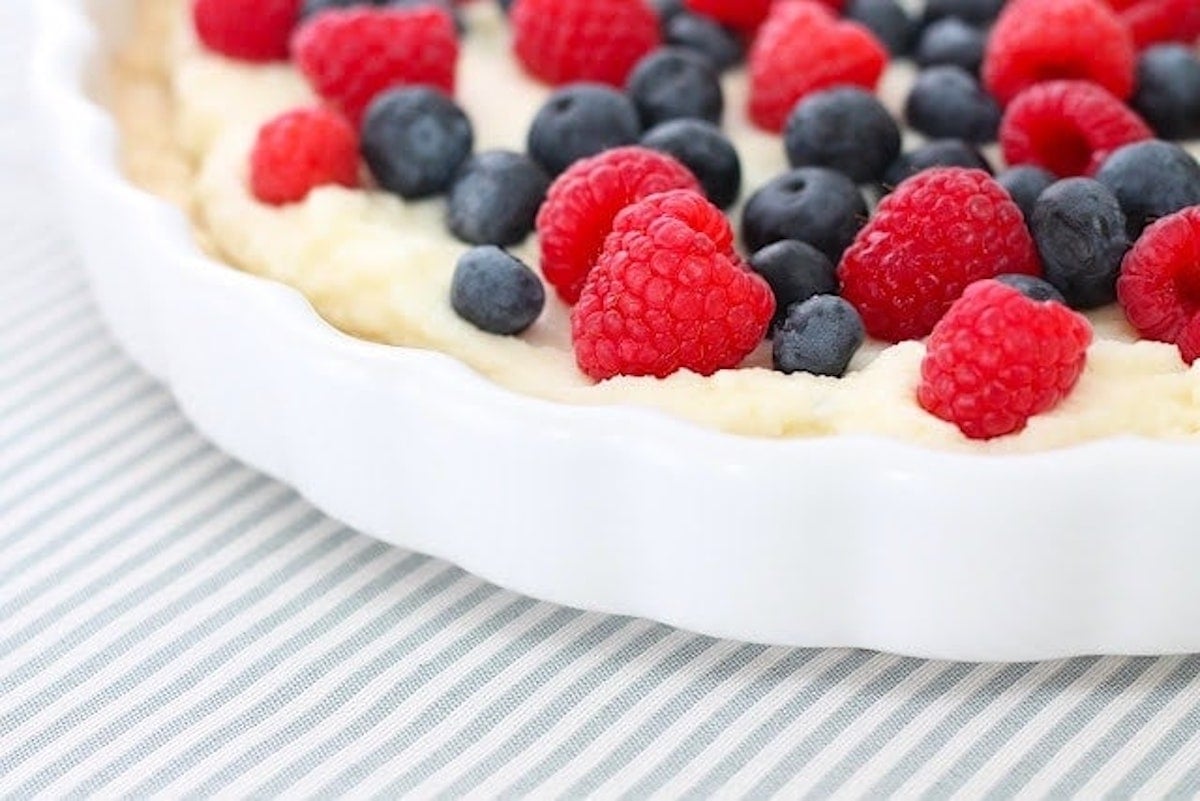 Close-up of a lemon berry tart in a white dish, topped with fresh raspberries and blueberries, placed on a light-colored, striped fabric surface.