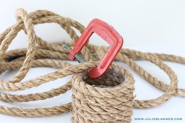 A clamp holding down the rope on a glass vase that is being being wrapped in it, on a white surface. 