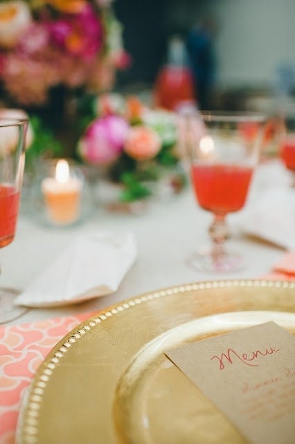 A close up a gold plate with of a pink, orange, green, and purple flowers with a raspberry cocktail to the side in the background.