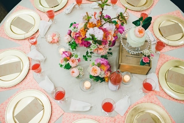 A set table with gold plates, pink, orange, green, and purple flowers with a raspberry cocktail to the side.