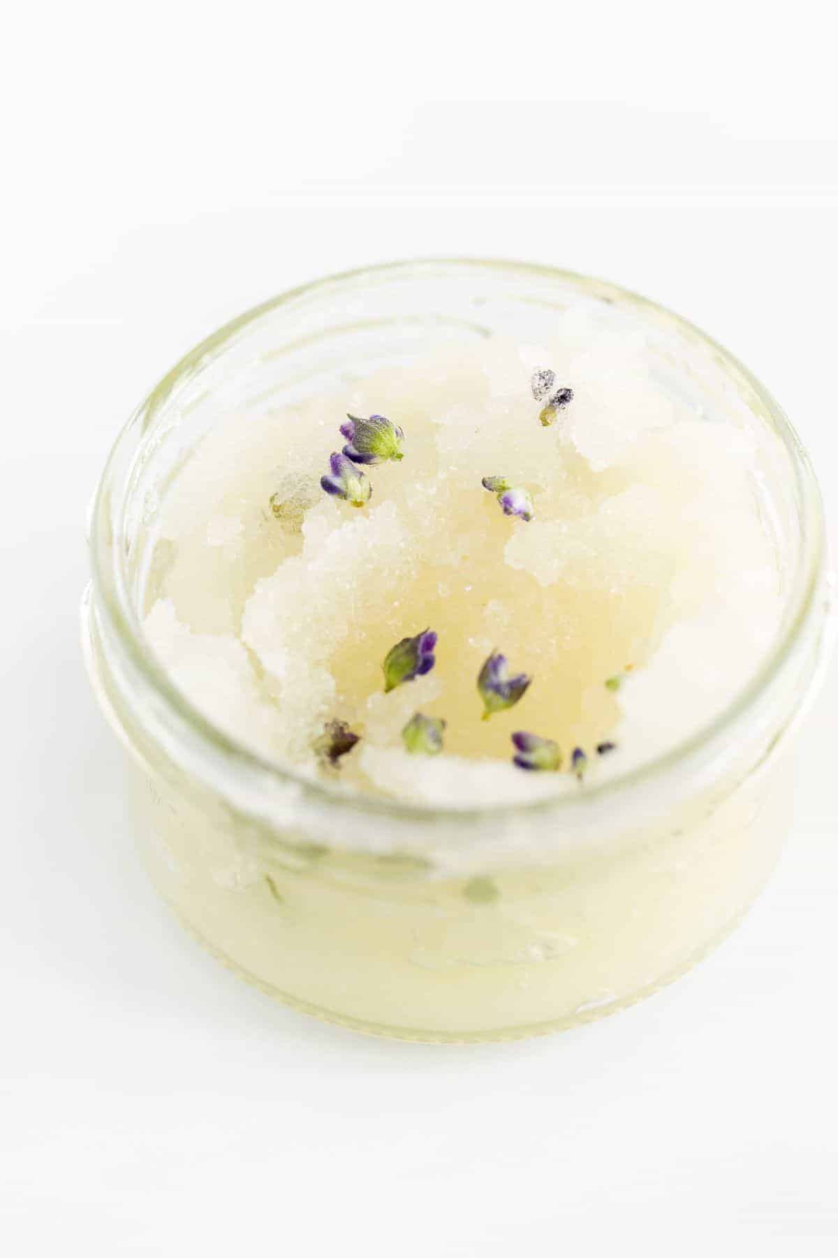 A homemade sugar scrub with lavender, in a glass jar on a white surface.