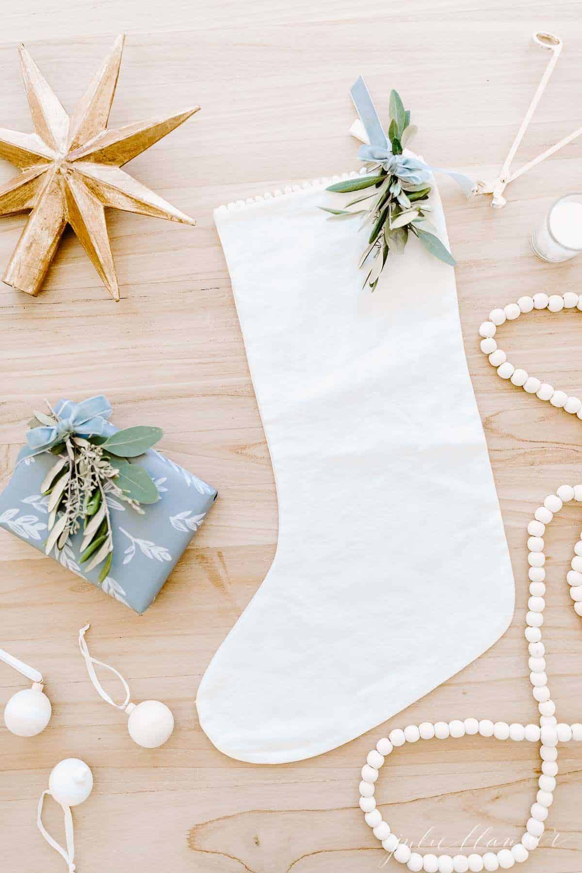 A wooden surface with a linen stocking, a gift card in a blue wrapped box, and wooden Christmas ornaments surrounding.