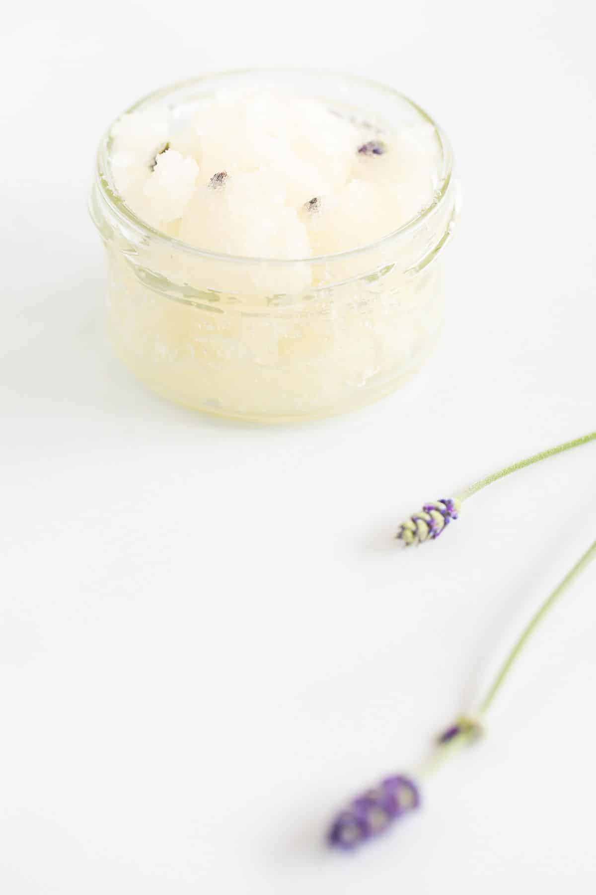 A homemade sugar scrub with lavender, in a glass jar on a white surface.