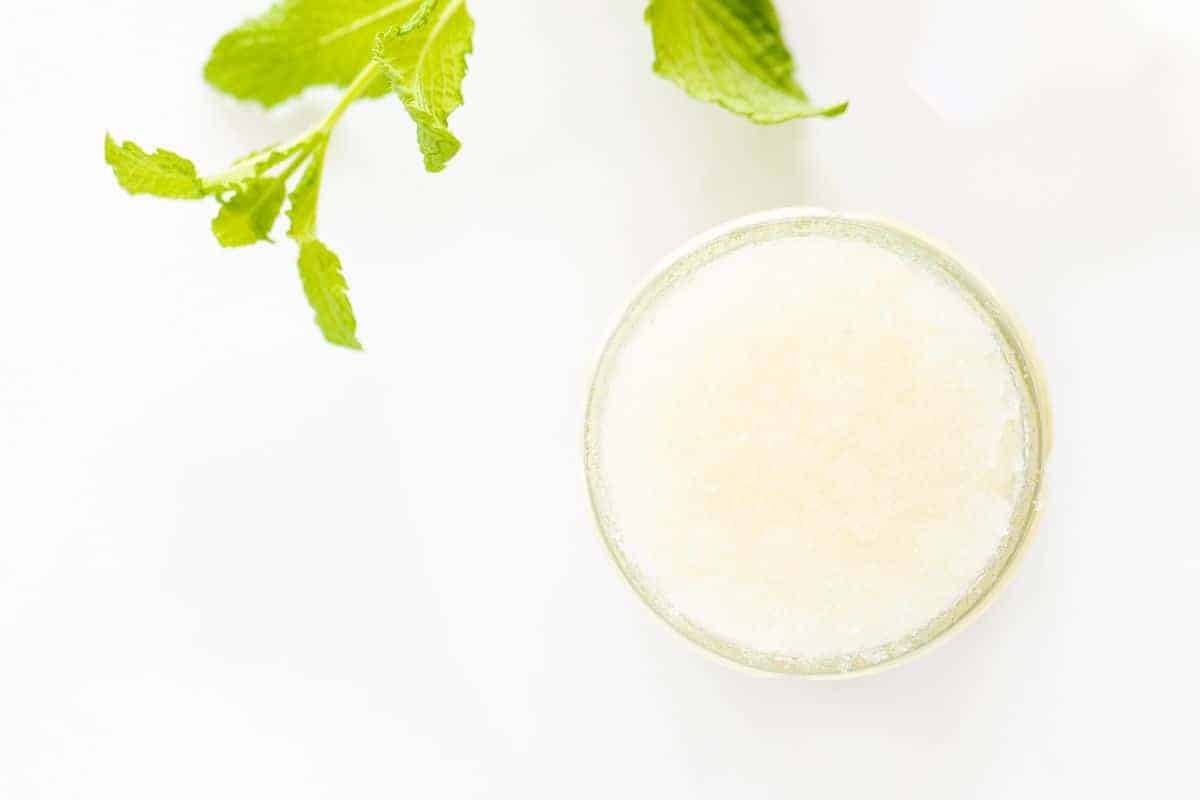 A glass jar full of a sugar scrub recipe, stem of fresh mint nearby.