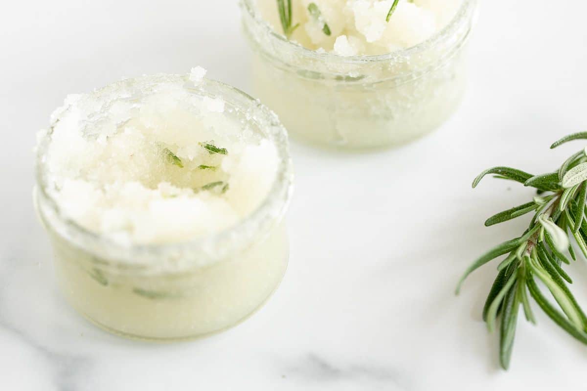 Two jars of homemade sugar scrub on a marble surface, stem of fresh rosemary in foreground.