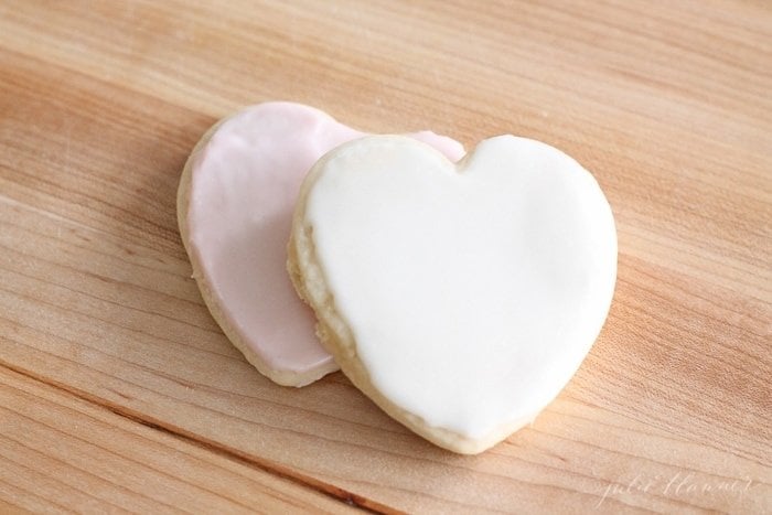 Two heart shaped cookies topped with sugar cookie icing on a wooden surface. 