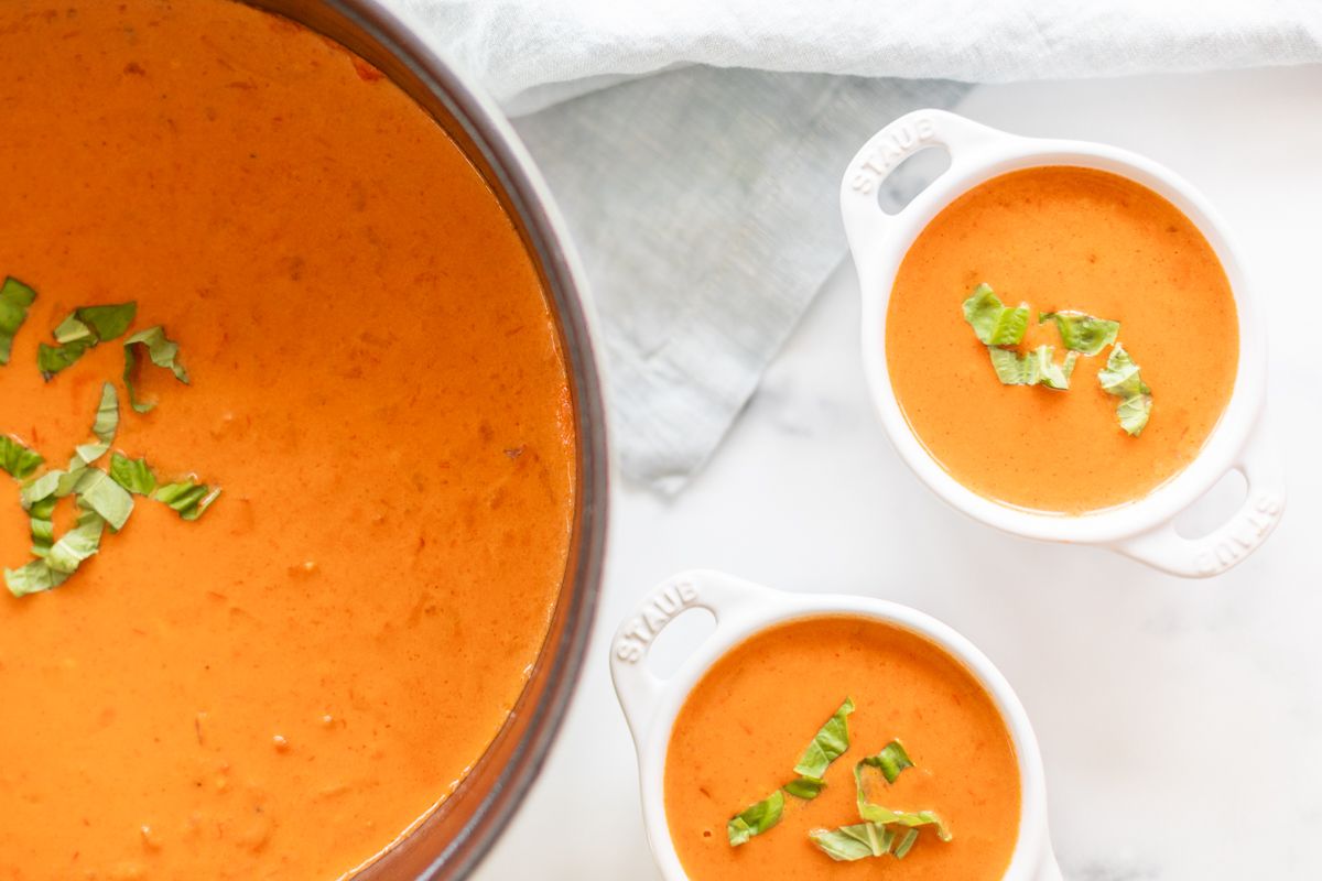 A cast iron pot filled with creamy tomato soup, garnished with fresh basil. Bowls of soup to the side.