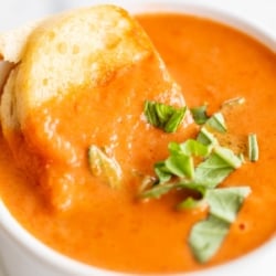 A white bowl full of a creamy tomato soup recipe, garnished with fresh basil. A Small grilled cheese sandwich dunks into the corner of the bowl.