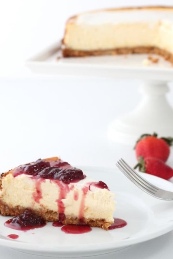 A slice of New York cheesecake on a white plate, with the whole cheesecake on a white cake pedestal in the background.