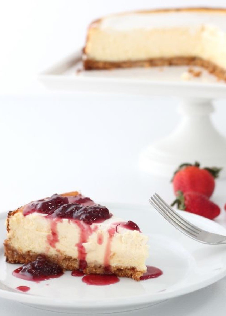 A slice of New York cheesecake on a white plate, with the whole cheesecake on a white cake pedestal in the background.