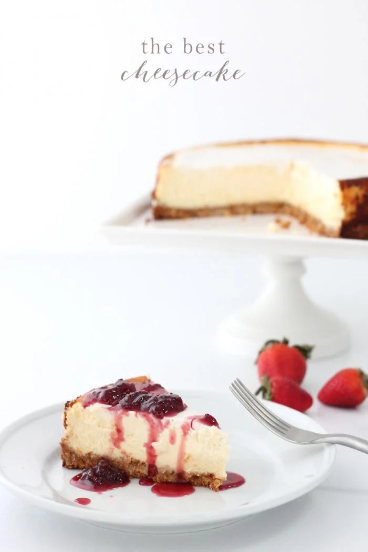 A slice of New York cheesecake on a white plate, with the whole cheesecake on a white cake pedestal in the background.