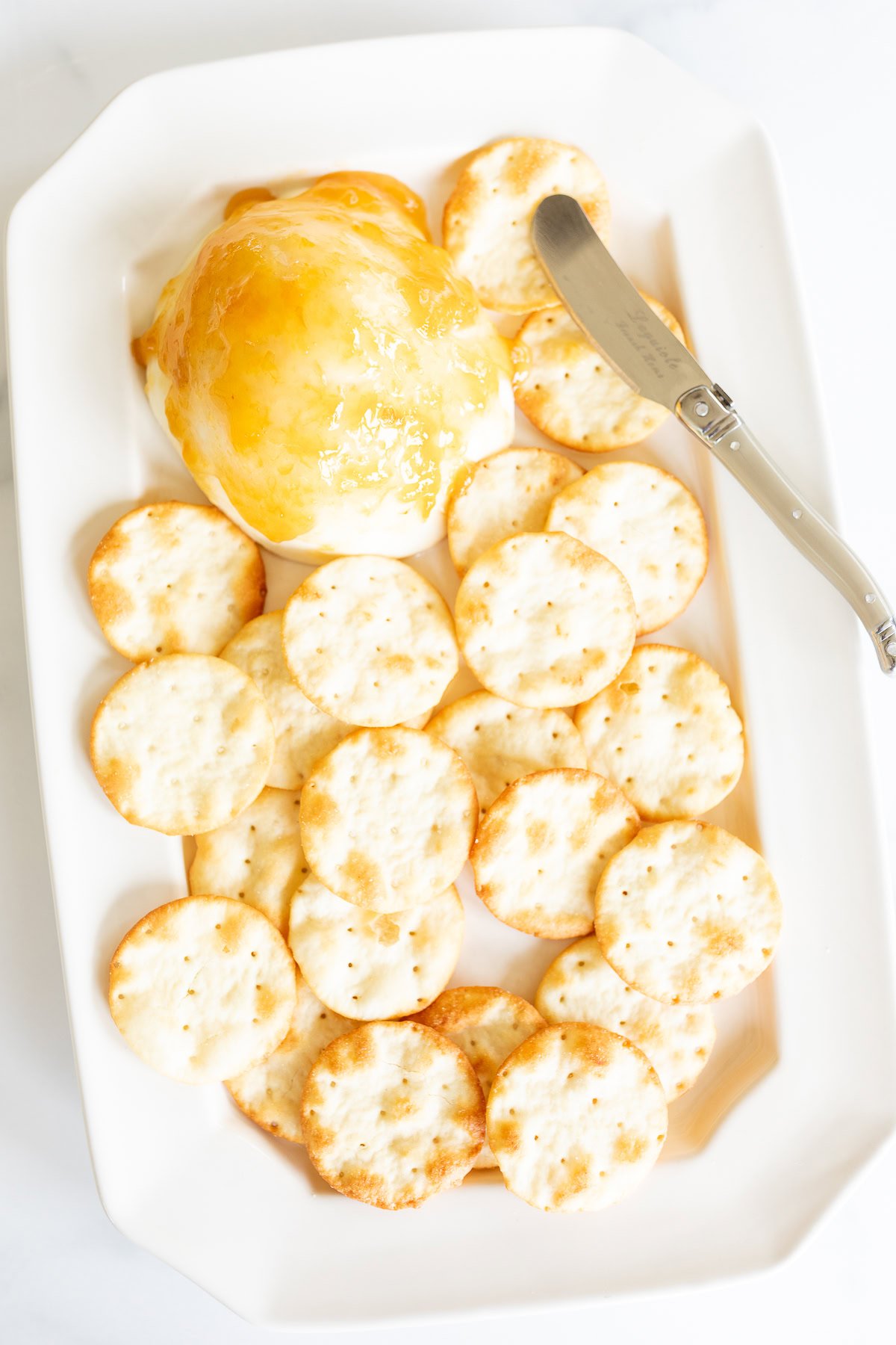 Grilled Mozzarella appetizer with apricot jam on top served on a white plate with crackers arranged around it, accompanied by a cheese knife.