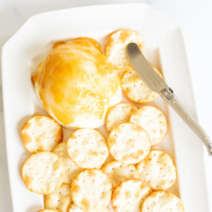 A close-up of a grilled mozzarella cheese ball topped with apricot jam, served with a spreader knife and surrounded by round crackers.