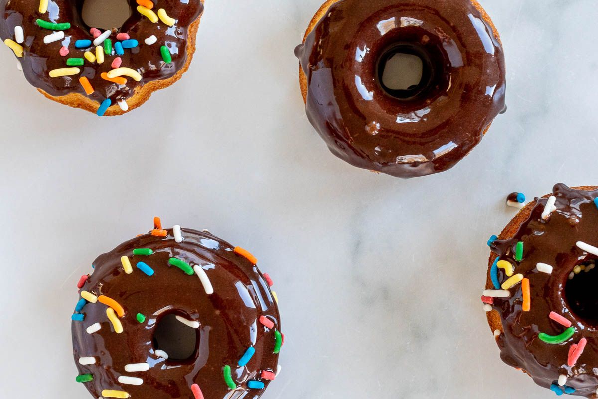 Baked donuts topped with chocolate frosting and sprinkles, on a marble countertop.