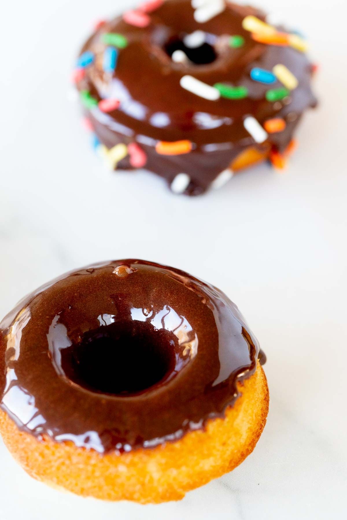 Baked donuts topped with chocolate frosting and sprinkles, on a marble countertop.