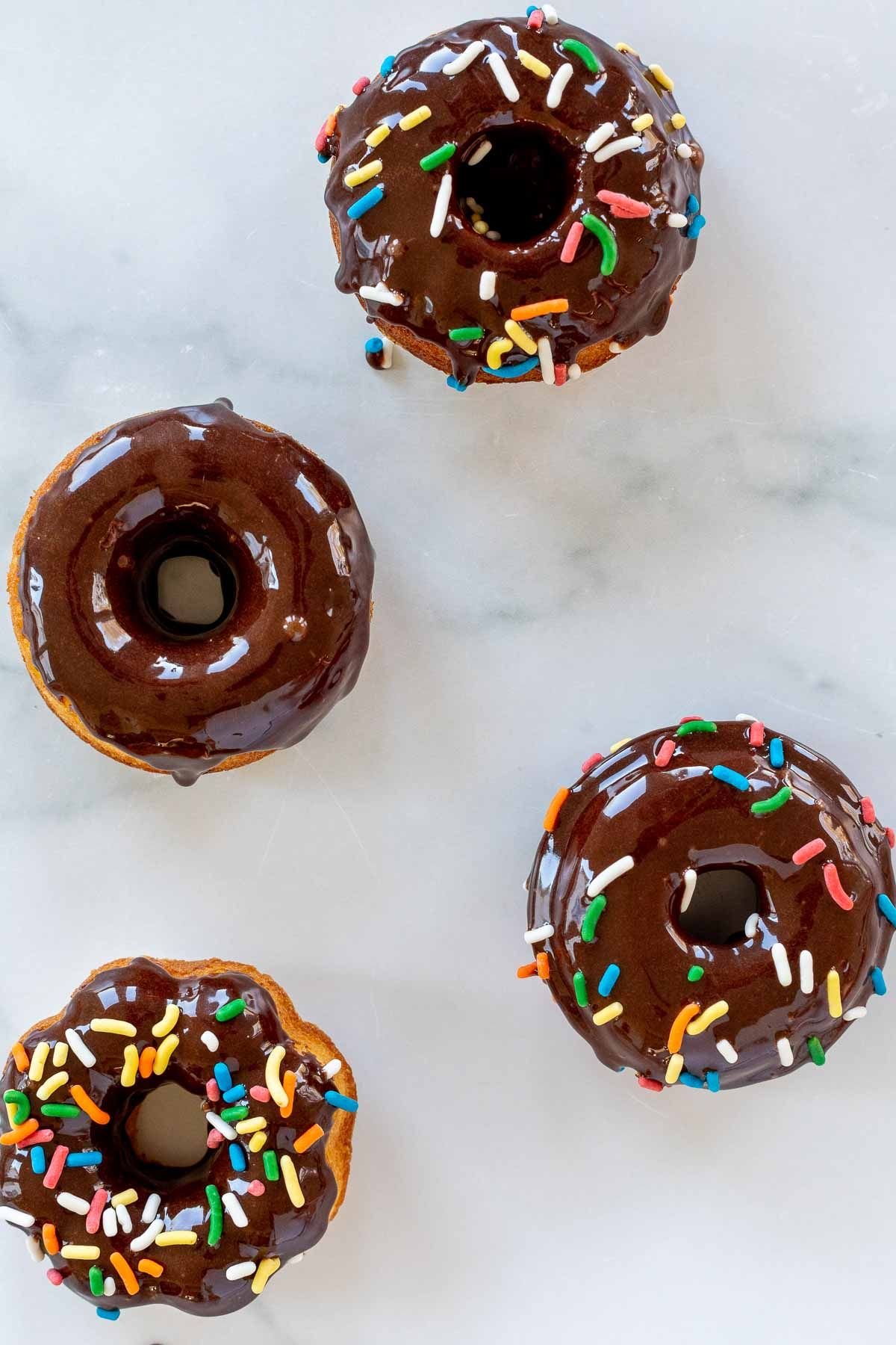 Baked donuts topped with chocolate frosting and sprinkles, on a marble countertop.