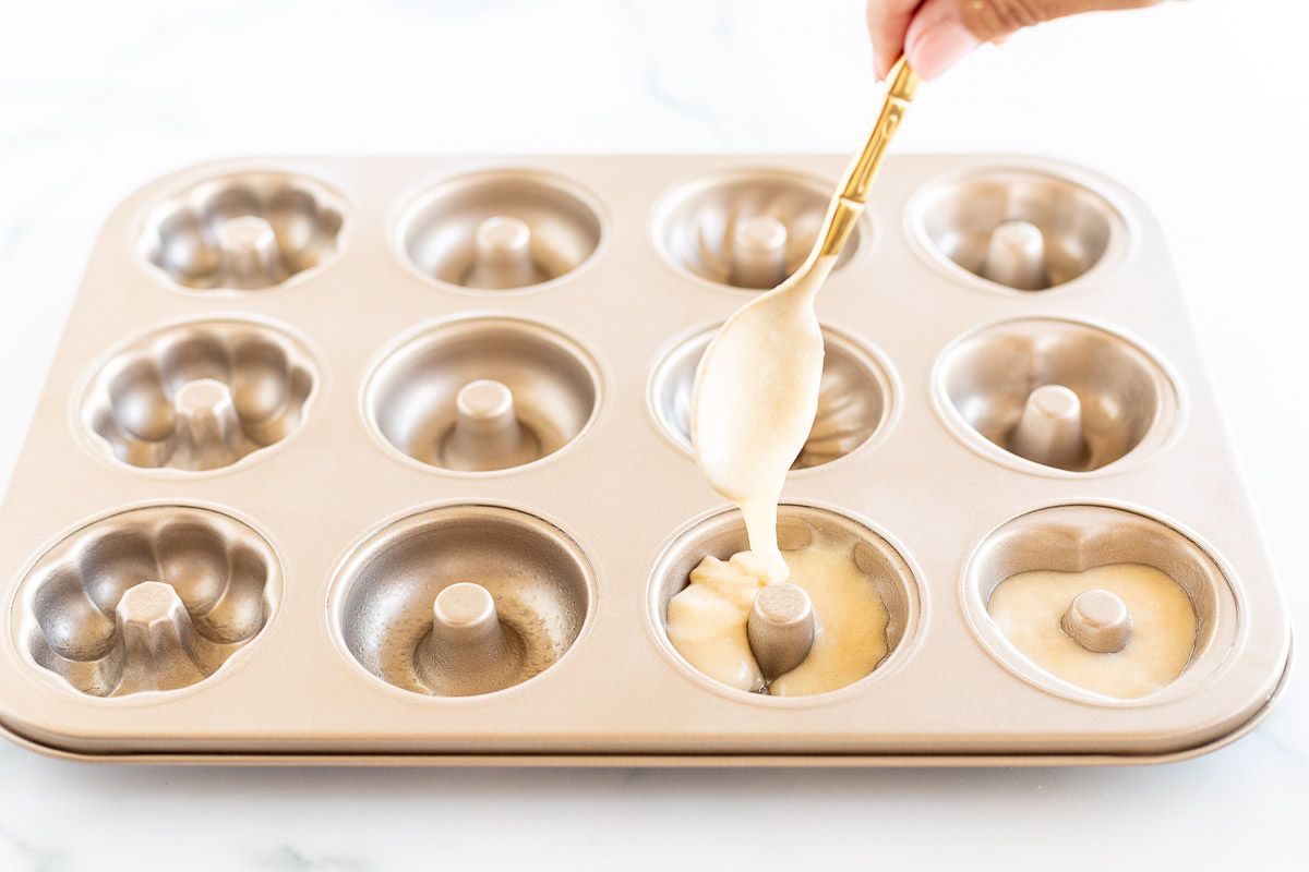 A hand spooning baked donut batter into a gold donut pan. 