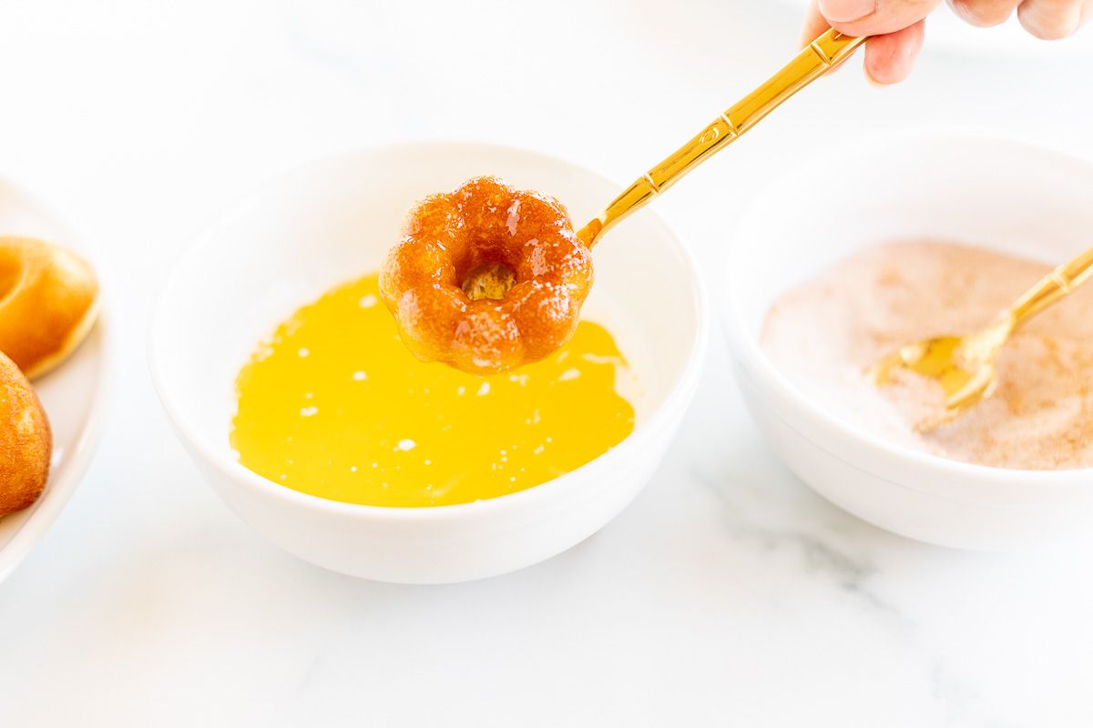 A baked donut being dipped into butter and a cinnamon sugar mixture.