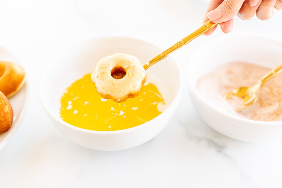 A baked donut being dipped into butter and a cinnamon sugar mixture.