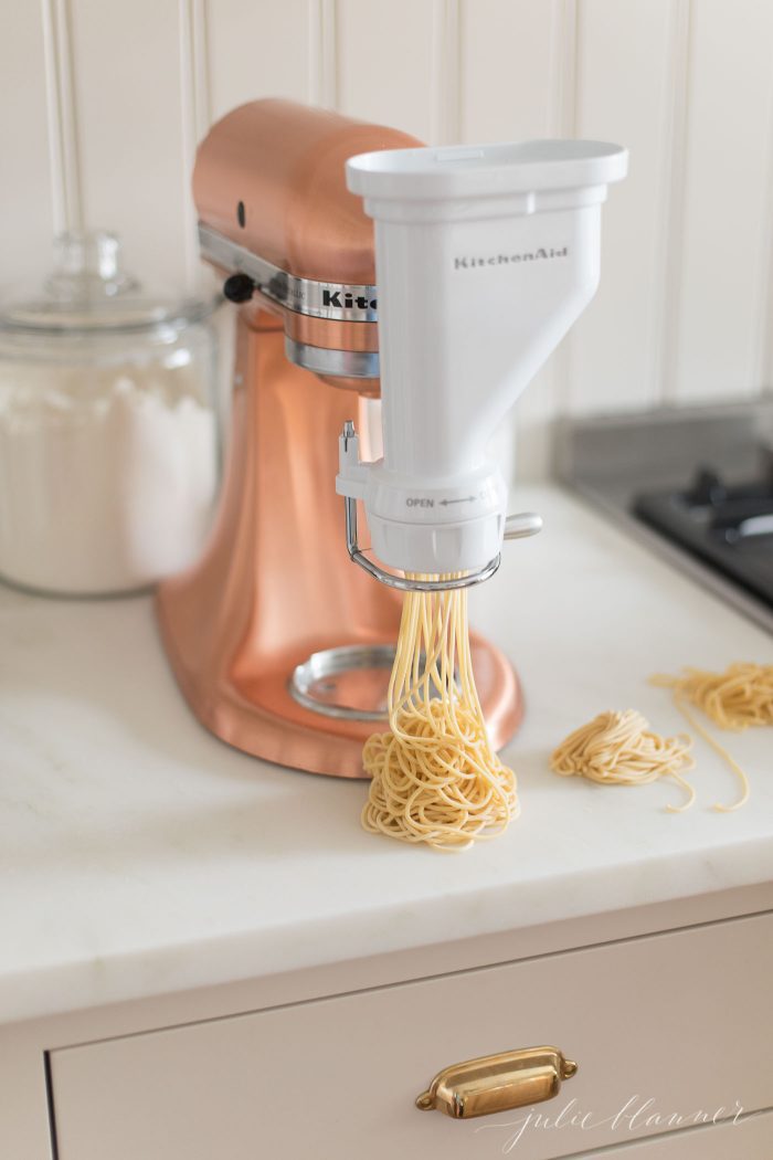 A copper Kitchenaid mixer with a pasta attachment. A small pile of spaghetti shaped egg noodles are piling onto a marble countertop.