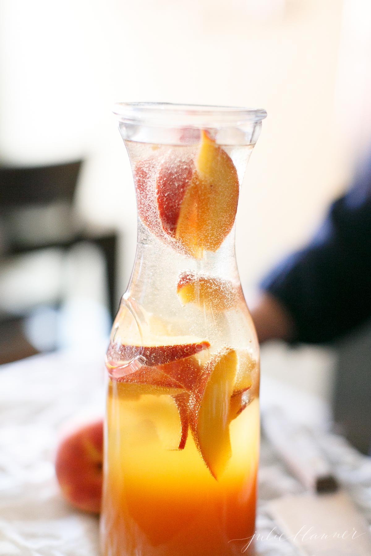 Prosecco Sangria in a glass carafe with pomegranate and peaches in the back.