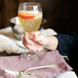 Prosecco Sangria in a glass carafe with pomegranate and peaches in the back.