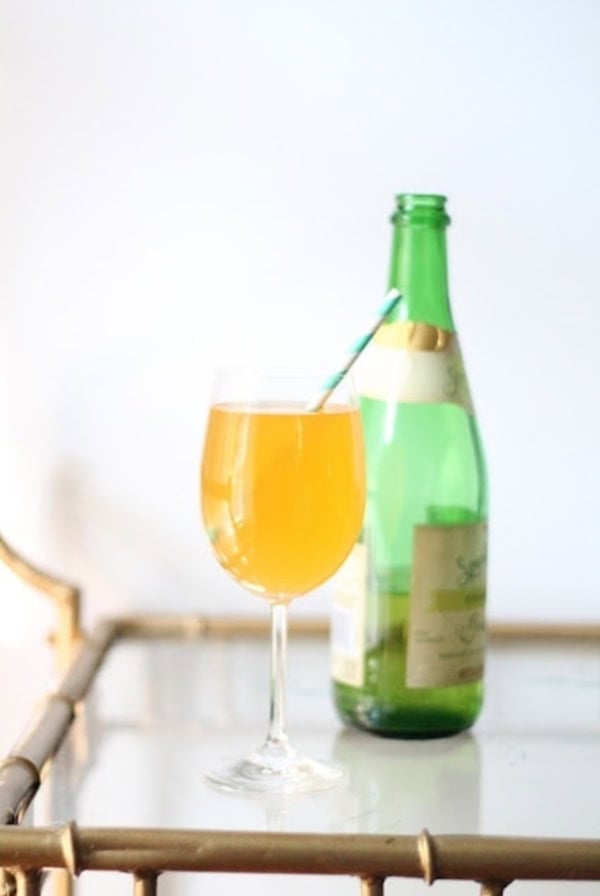 A green glass bottle next to a wine glass filled with an orange-yellow Non-Alcoholic Bellini sits elegantly on a glass and wood table, perfect for a baby shower cocktail.