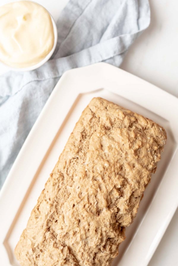 A loaf of beer bread mix on a white rectangular plate with a light blue cloth napkin in the background and a small bowl of creamy spread beside it.