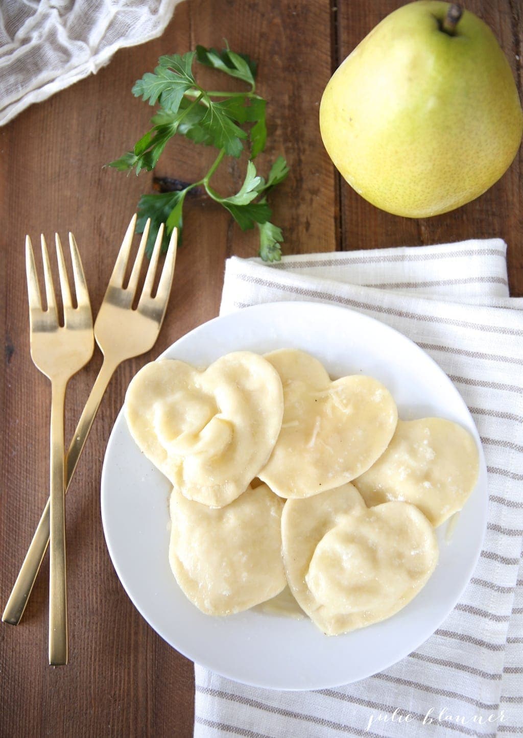 heart shaped ravioli on a white plate, gold forks to the side and a pear in the background.