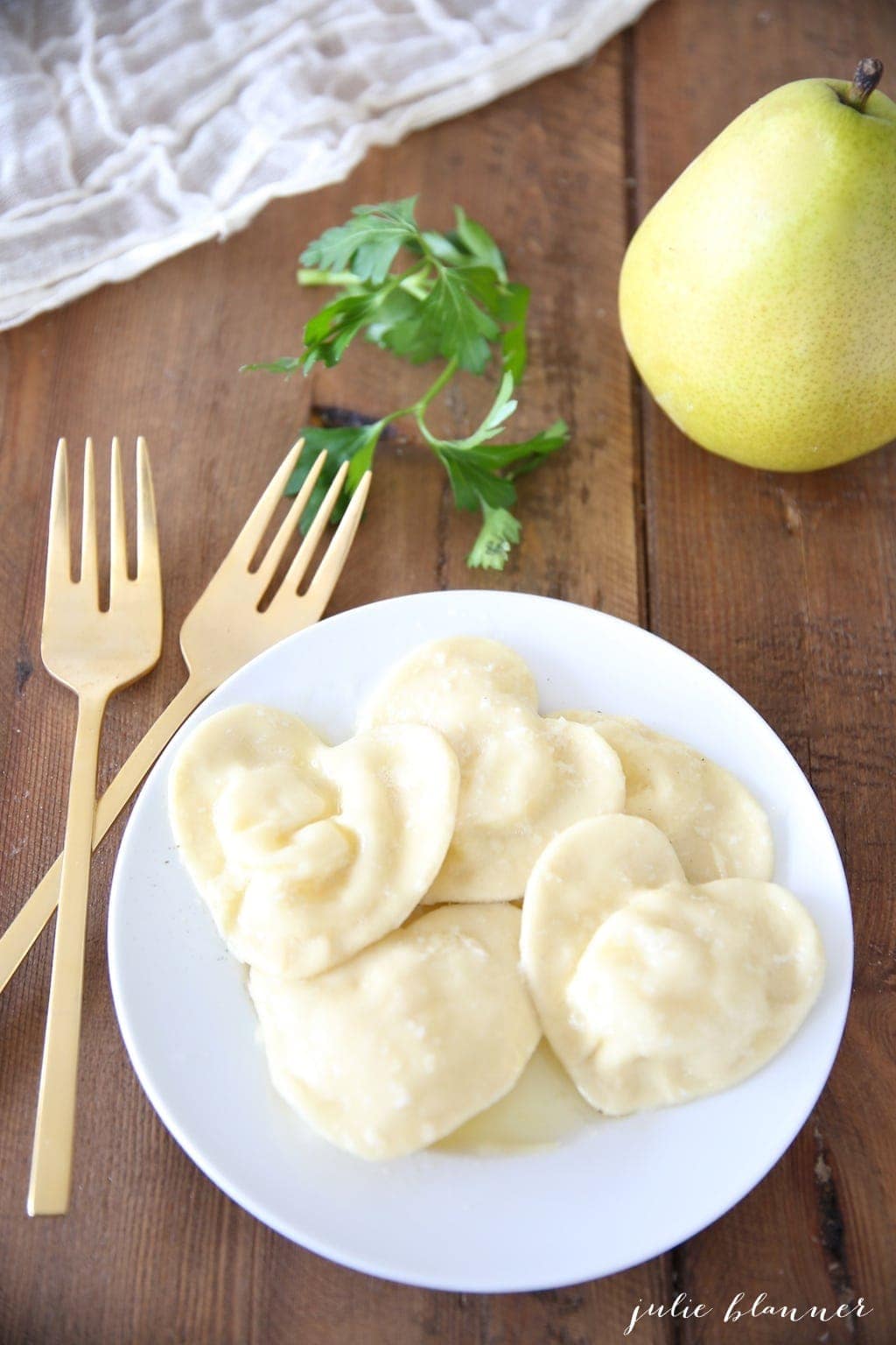 heart shaped ravioli on a white plate, gold forks to the side and a pear in the background.