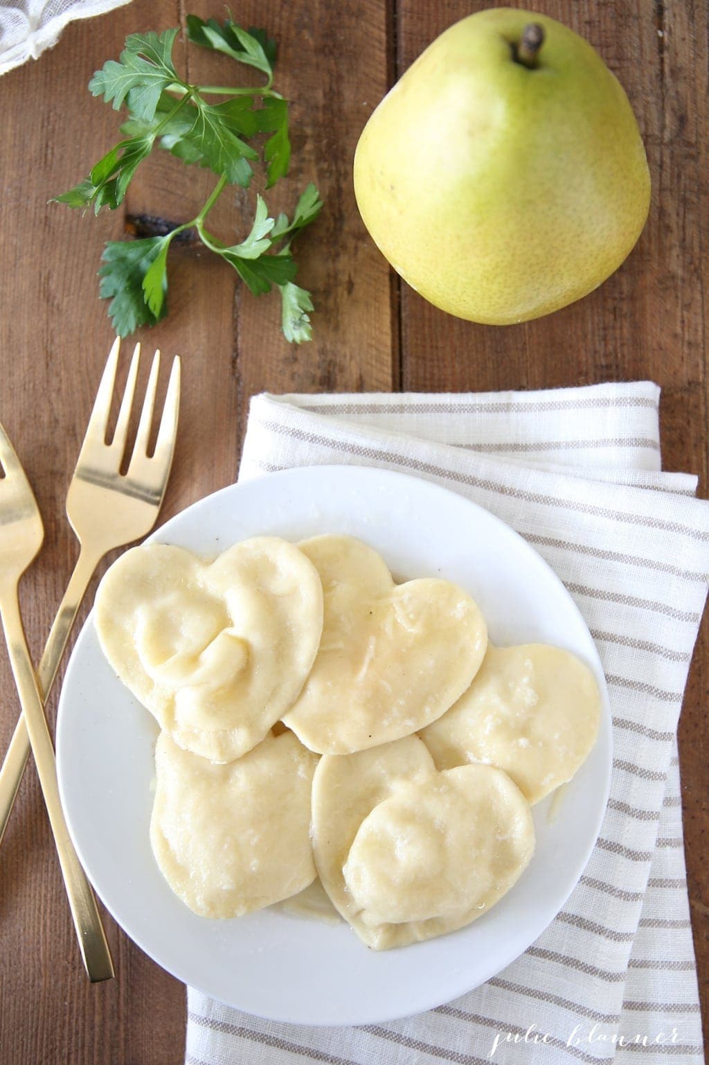 heart shaped ravioli on a white plate, gold forks to the side and a pear in the background.