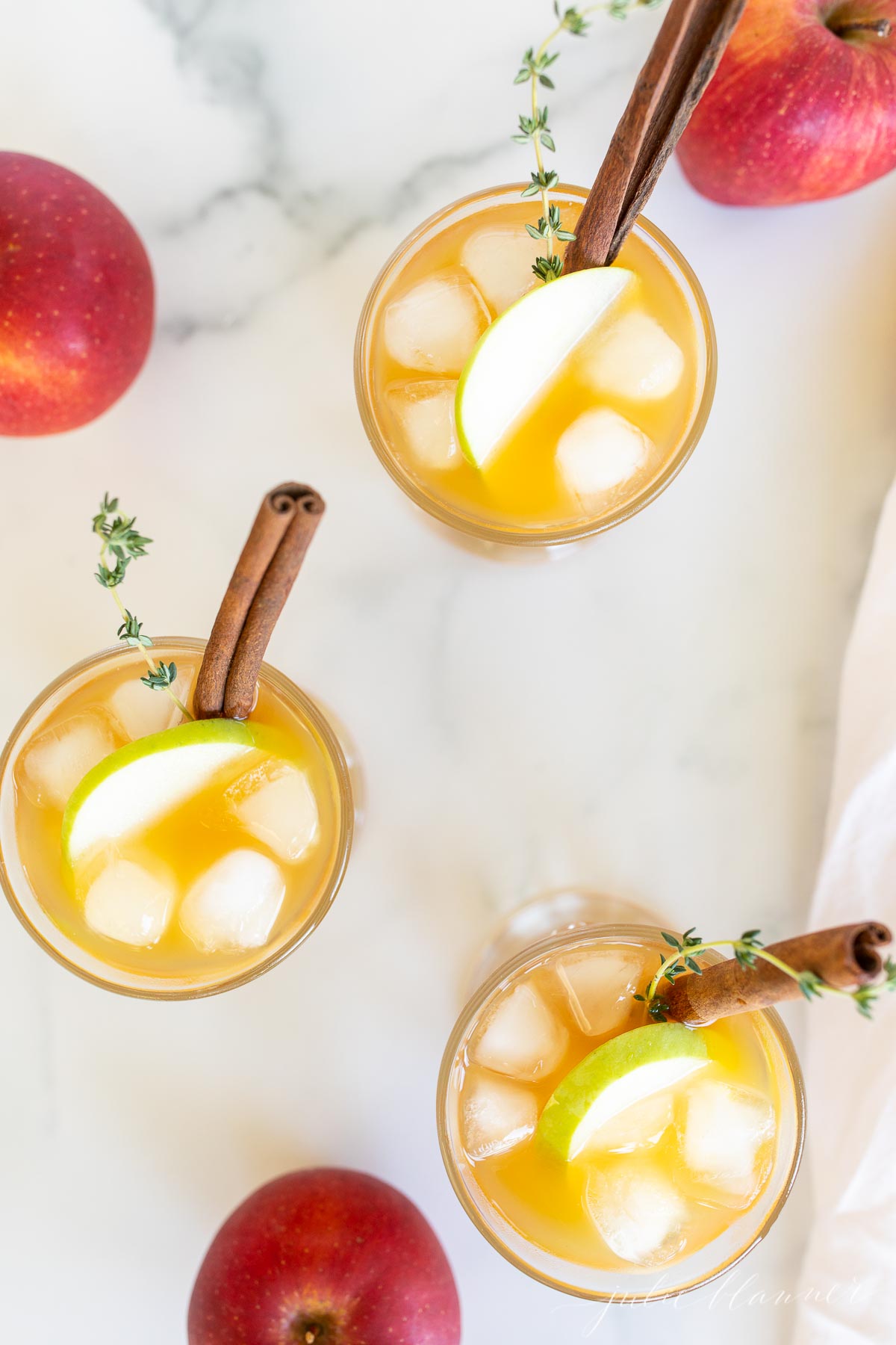 Three glasses filled with apple cider cocktails, garnished with apple slices and a cinnamon stick.