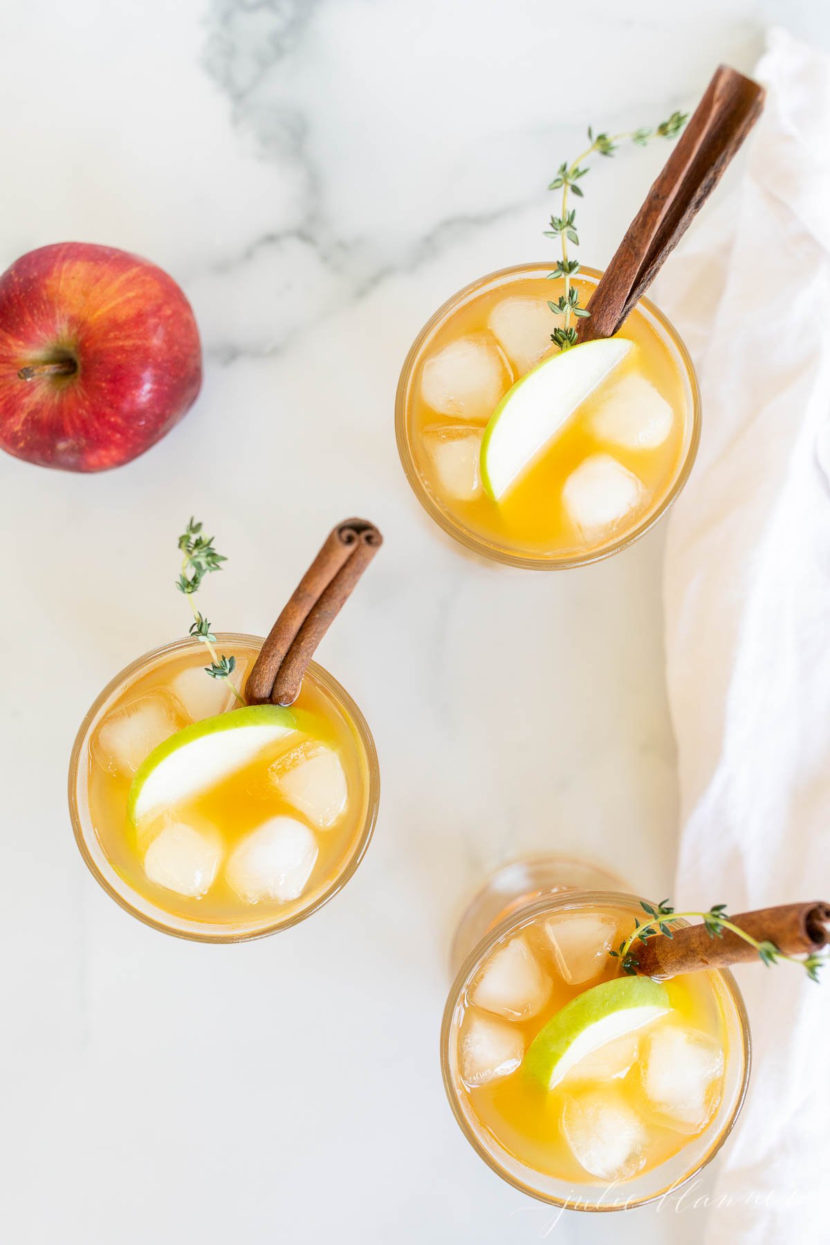 Three glasses filled with apple cider cocktails, garnished with apple slices and a cinnamon stick.