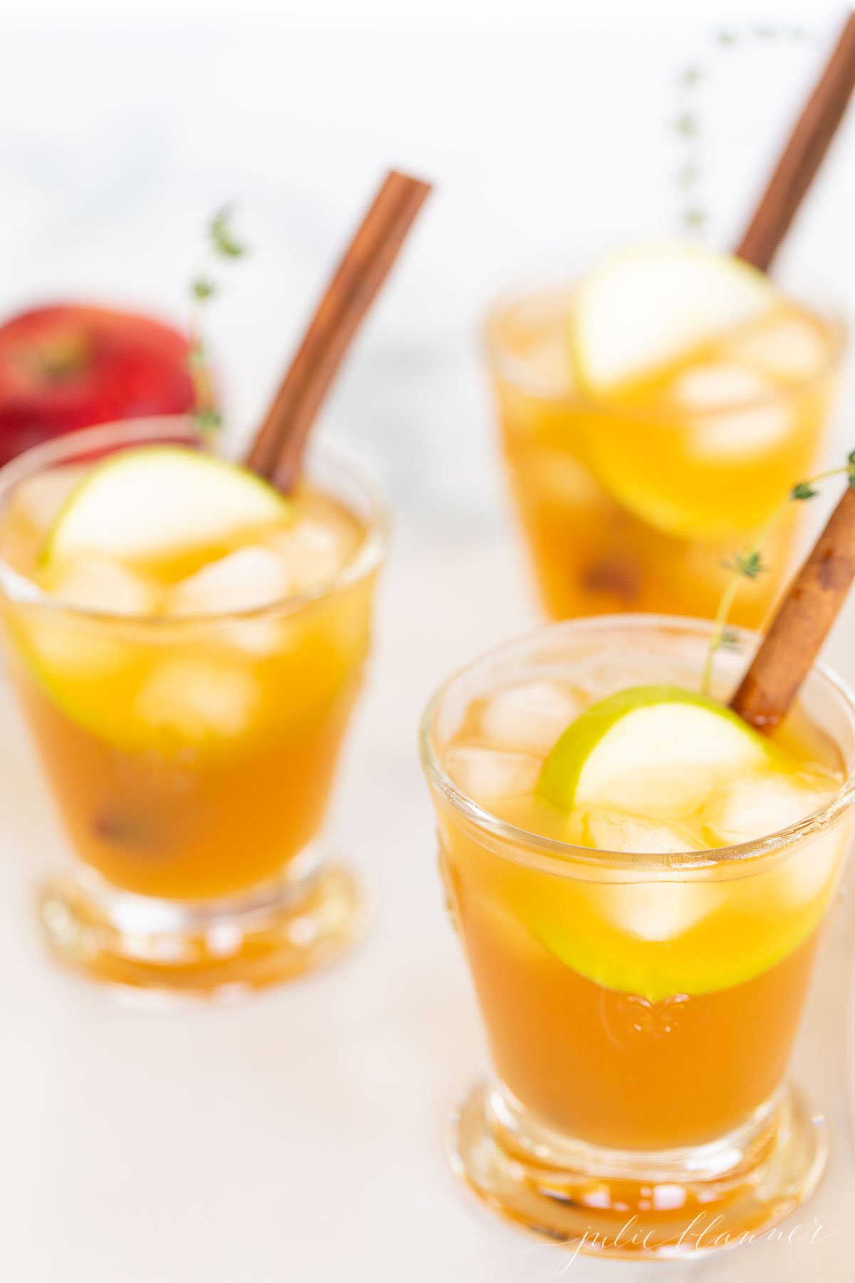 Three glasses filled with apple cider cocktails, garnished with apple slices and a cinnamon stick.