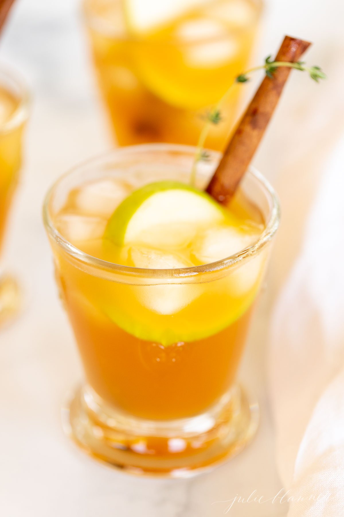 Three glasses filled with apple cider cocktails, garnished with apple slices and a cinnamon stick.