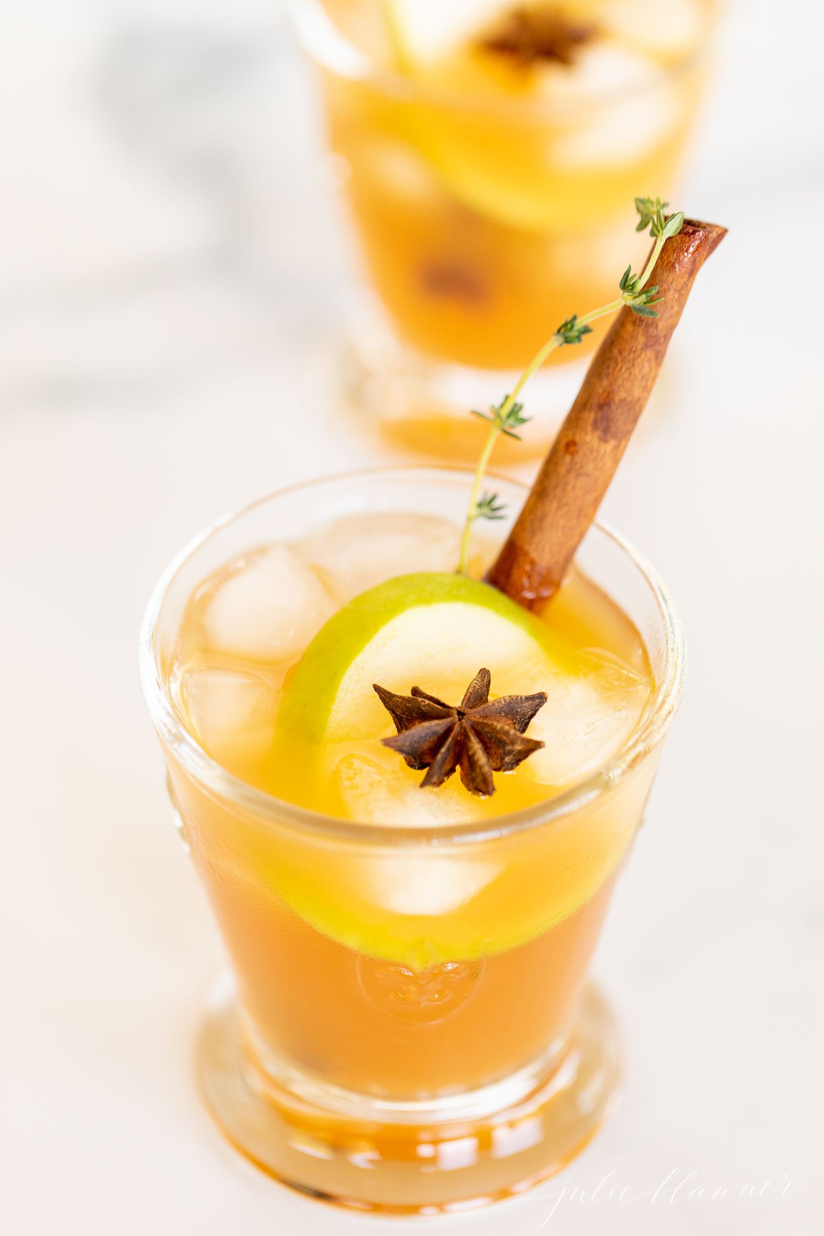 An apple cider cocktail on a white marble countertop, garnished with an apple slice and cinnamon stick.